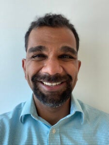 Sandesara, a middle aged South Asian man with black short curly hair and a salt and pepper beard, smiles into the camera. He is wearing a checkered blue button up.